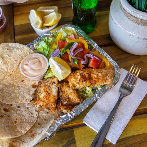 Grilled fish, salad, tortillas, and sauce in a foil container, with lemon wedges and a drink.