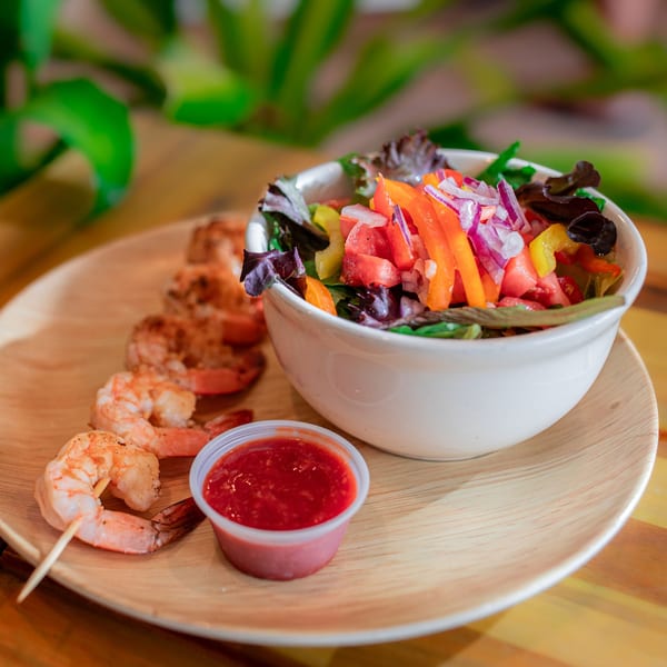Grilled shrimp skewers, a side salad with colorful vegetables, and a cup of red sauce on a wooden plate.