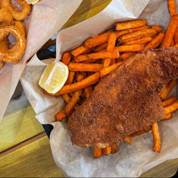 Crispy fried fish with sweet potato fries and a lemon wedge, served in a paper-lined basket.