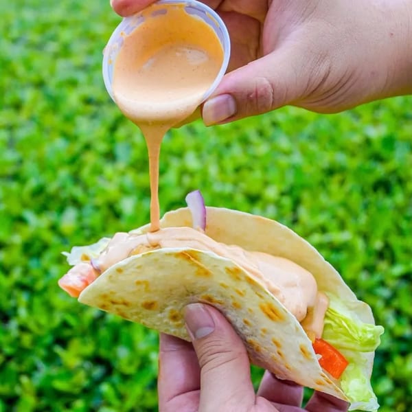 Hands pouring creamy sauce from a small cup onto a fish taco with lettuce and red onion.