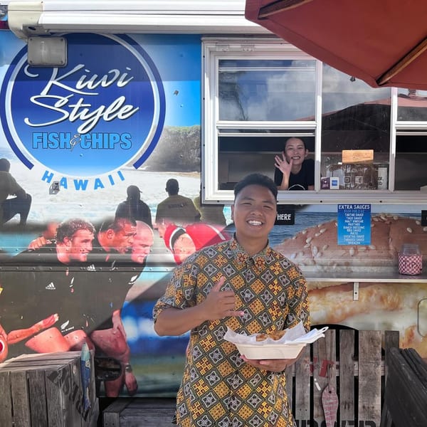 A man holds a tray of Kiwi Style Fish and Chips in front of a food truck, with a woman waving from inside.
