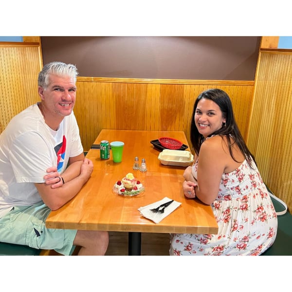 A smiling couple sits at a wooden table with a dessert, drinks, and takeout container.