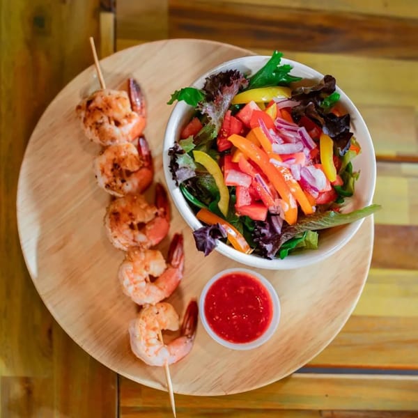 Grilled shrimp skewers, a fresh garden salad, and a small bowl of red dipping sauce on a wooden platter.