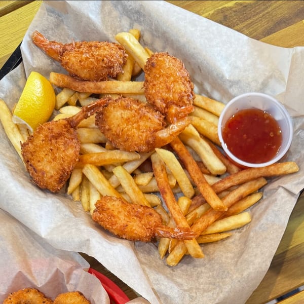 A basket of golden-brown coconut shrimp and french fries with a lemon wedge and sweet chili sauce.