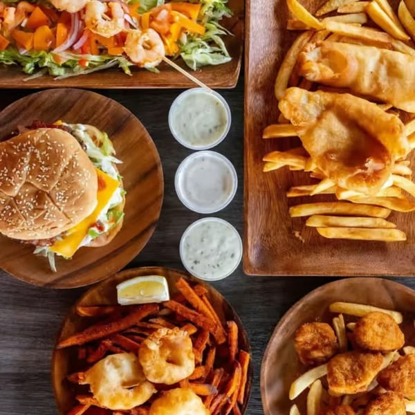Overhead view of various fried seafood dishes, a burger, and fries on wooden plates with dipping sauces.