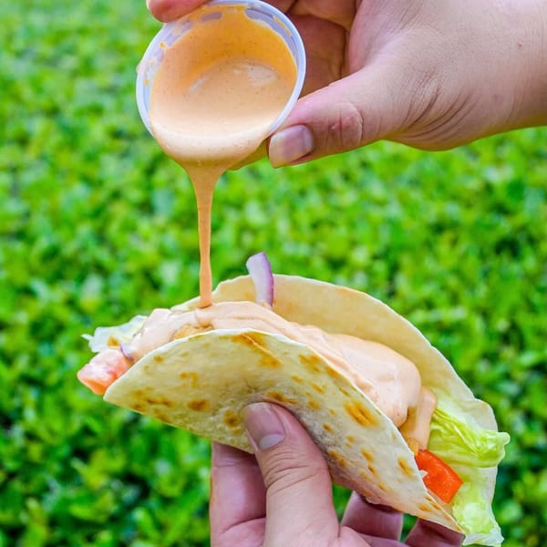 Hands pouring creamy sauce from a cup onto a taco filled with lettuce, red peppers, and onions.