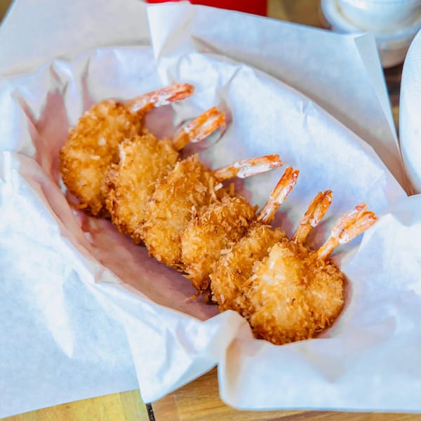Five crispy, golden-brown coconut shrimp with tails on, served in a paper-lined basket.