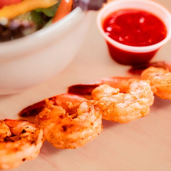 Grilled shrimp with a side of red dipping sauce and a salad in a white bowl.